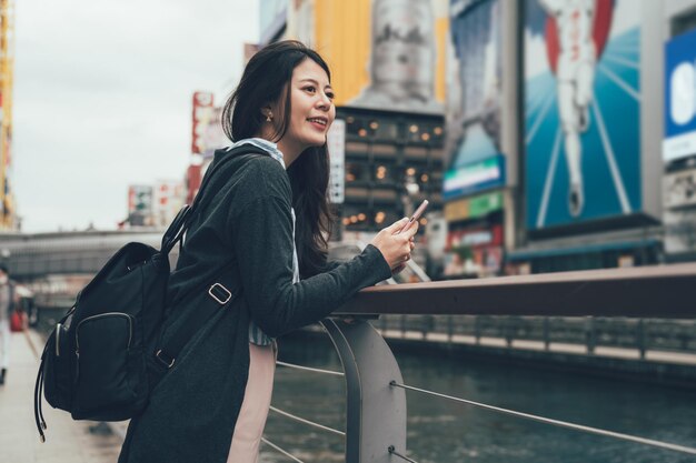 Una mochilera alegre y guapa sostiene un teléfono móvil mientras se apoya en las barandillas del puente y mira disfrutar de la hermosa vista del canal del río en dotonbori. nippon runner running man cartel publicitario.