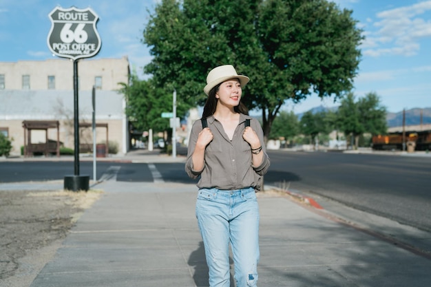 una mochilera asiática agarrando correas está explorando una ciudad rural en los EE. UU. una viajera coreana con sombrero de navegante paseando por la carretera de un estado con un cartel y mirando alrededor de la zona.