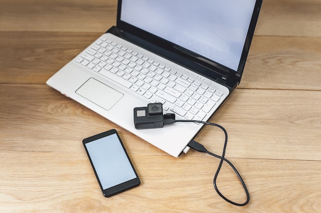 Mock-se de um smartphone e uma câmera de ação conectada a um laptop branco sobre um fundo de mesa de madeira.