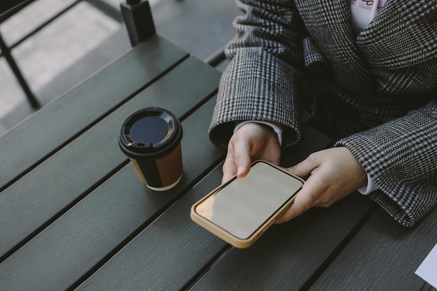 Mock-up-Bild von weiblichen Händen, die Handy mit leerem Bildschirm und Tasse Kaffee in einem Café auf der Straße halten.