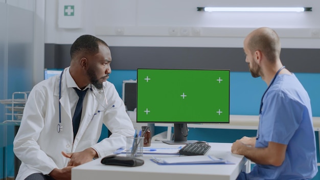 Mock-up-Chroma-Key-Computer mit grünem Bildschirm mit isolierter Anzeige auf dem Tisch im Krankenhausbüro. Multiethnisches medizinisches Team, das Krankheitssymptome diskutiert, die in der Gesundheitsversorgung arbeiten
