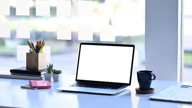 Mock-up-Laptop-Computer mit weißem Bildschirm, Kaffeetasse und Bürobedarf auf weißem Tisch.