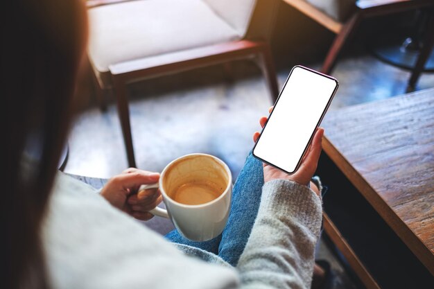 Mockup-Bild einer Frau, die beim Kaffeetrinken ein Mobiltelefon mit einem leeren weißen Desktop-Bildschirm hält