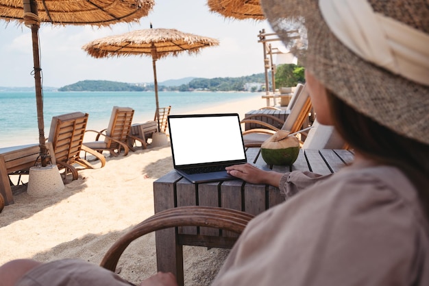 Mockup-Bild einer Frau, die ein Laptop-Touchpad mit leerem Desktop-Bildschirm verwendet und berührt, während sie am Strand sitzt