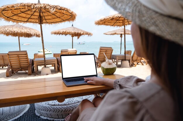 Mockup-Bild einer Frau, die ein Laptop-Touchpad mit leerem Desktop-Bildschirm verwendet und berührt, während sie am Strand sitzt