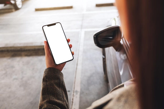 Mockup-Bild einer Frau, die ein Mobiltelefon mit einem leeren weißen Bildschirm neben dem Auto auf dem Parkplatz hält und benutzt