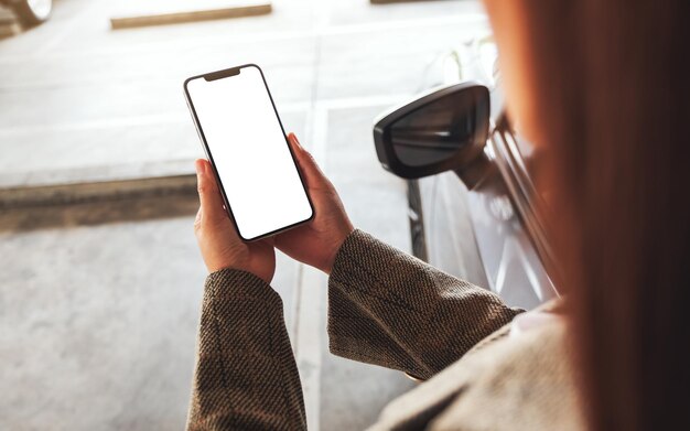 Mockup-Bild einer Frau, die ein Mobiltelefon mit einem leeren weißen Bildschirm neben dem Auto auf dem Parkplatz hält und benutzt