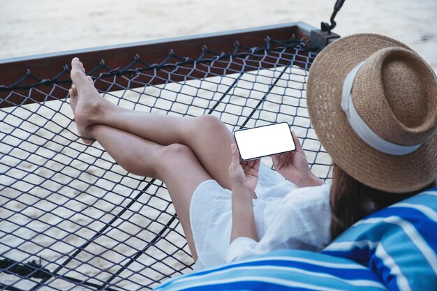 Mockup-Bild einer Frau, die ein Mobiltelefon mit leerem Desktop-Bildschirm hält, während sie sich auf einer Hängematte am Strand hinlegt