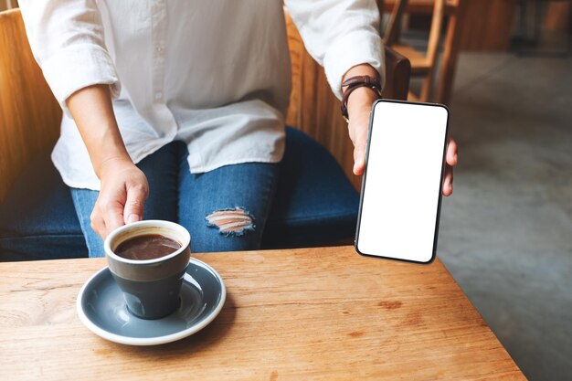 Mockup-Bild einer Frau, die ein Mobiltelefon mit leerem weißem Bildschirm hält und zeigt, während sie im Café Kaffee trinkt