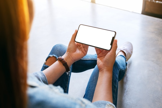 Mockup-Bild einer Frau, die ein schwarzes Handy mit leerem weißem Bildschirm hält, während sie auf dem Boden sitzt