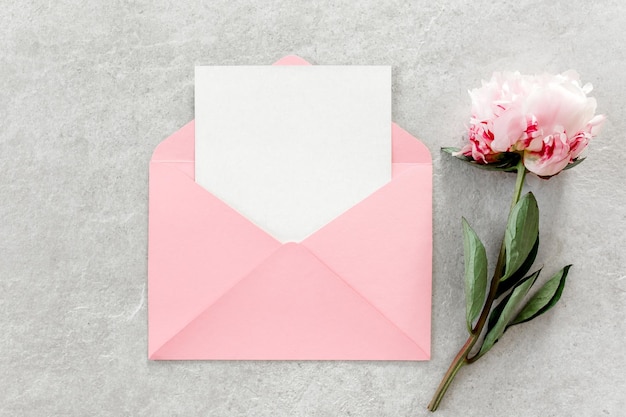 Mockup de convite papel em branco cartão de saudação envelope rosa e peônias em mesa de pedra cinza fundo de flor vista superior plana