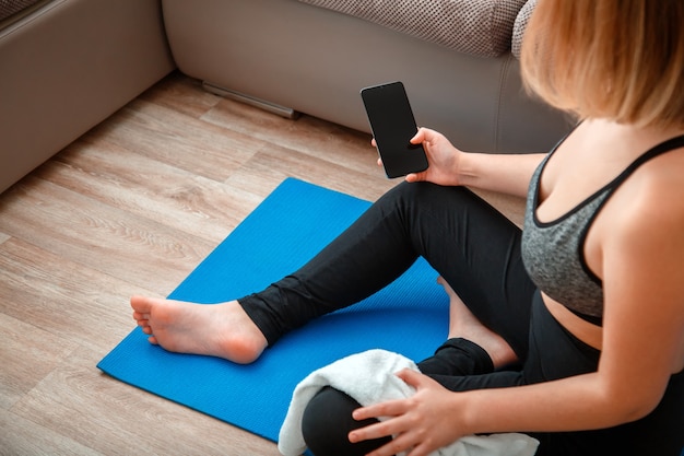 Mockup für App-Online-Fitnessprogramm, Remote-Yoga-Kurs. Frau in Sportbekleidung mit Smartphone mit leerem Bildschirm für Sport- oder Musik-App während einer Trainingspause auf der Matte zu Hause.