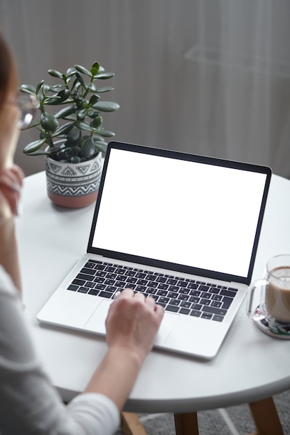 Mockup mulher de laptop de tela branca usando computador enquanto está sentado à mesa em casa vista traseira