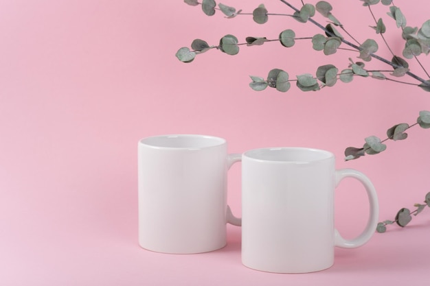 Mockup zwei weiße Kaffeetassen oder Becher auf rosafarbenem Hintergrund mit Kopierraum. Leere Vorlage für Ihr Design, Branding, Geschäft. Echtes Foto. Eukalyptuszweige