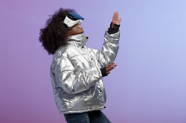 Mod chica de pelo castaño rizado vestida con una chaqueta de color plateado usa las gafas de realidad virtual en el estudio sobre fondo de neón.