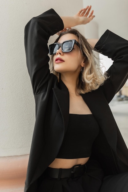 Moda adorable hermosa joven modelo de mujer elegante con elegantes gafas de sol en ropa de moda de negocios negra con blazer y top se sienta y posa en la ciudad cerca de una columna blanca vintage