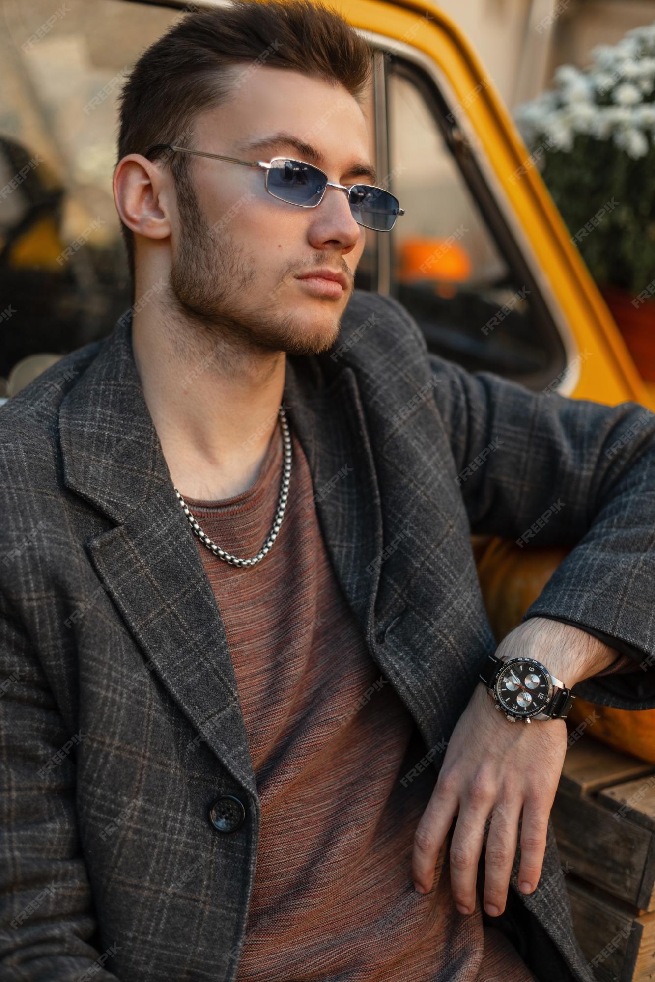 Moda apuesto hombre de negocios modelo hipster con elegantes gafas de sol  azules en un abrigo de moda con un elegante reloj de lujo se sienta cerca  de un coche retro amarillo