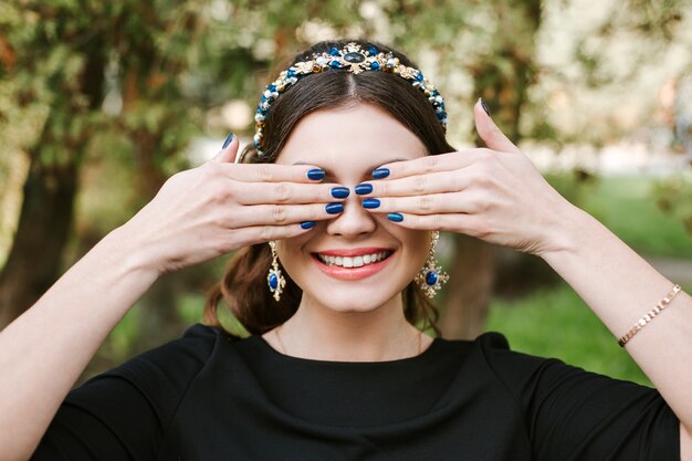 Moda, belleza, ternura, manicura. Mujer joven feliz con una brillante sonrisa de manicura amplia, sonrisa blanca, dientes rectos y blancos. La niña se cubre la cara con las manos. Cinta para el pelo, pendientes, esmalte de uñas azul.