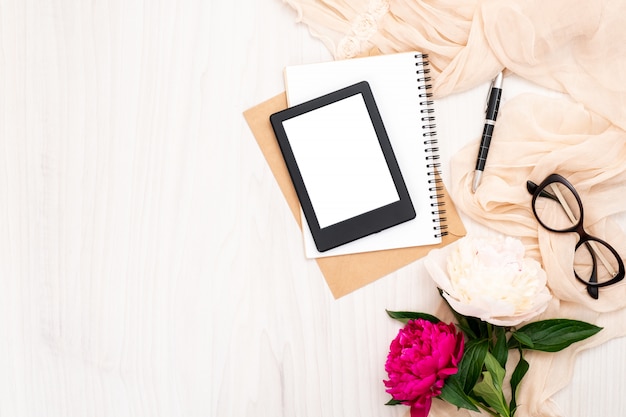 Moda blogger mesa de escritório em casa com itens de mulher: leitor de e-book moderno, bloco de notas de papel, cachecol bege, flores de peônias, óculos