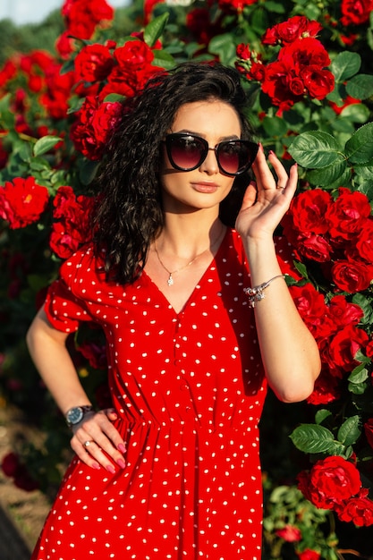 Moda bonita mulher com óculos de sol vintage e pulseira de joias em poses de vestido vermelho perto do arbusto com flores de rosas na luz do sol