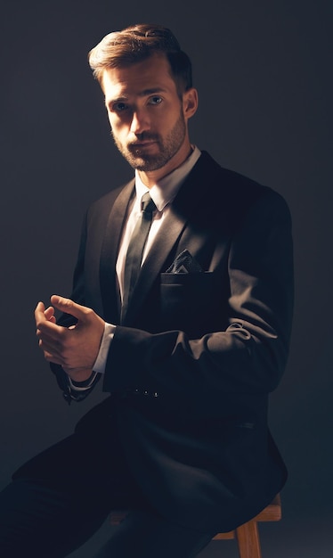 Moda de caballero y retrato de un hombre en traje con estilo para los negocios que parece serio en un fondo negro de estudio Modelo masculino seguro, orgulloso y guapo sentado en una silla ajustando las mangas