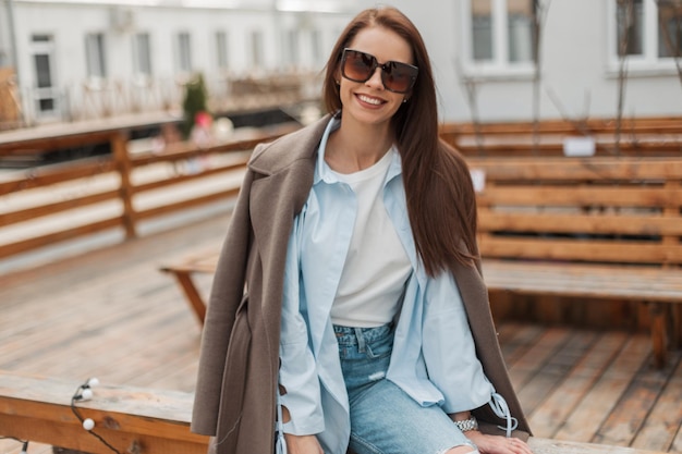 Moda casual jovem feliz com um sorriso doce com óculos de sol estilosos em roupas da moda com um casaco e jeans senta e se diverte na cidade