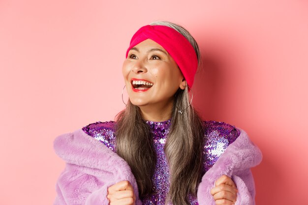 Moda. close-up de feliz mulher asiática sênior, sorrindo, olhando para a esquerda com uma cara alegre, em pé com um casaco de peles artificiais roxo, fundo rosa.