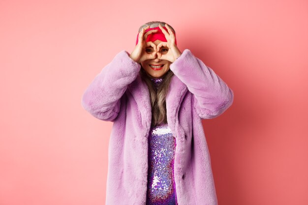 Moda y compras. Feliz abuela asiática en elegante abrigo de piel de faur, mirando a través de gafas de mano, haciendo binoculares y sonriendo a cámara, de pie sobre fondo rosa.