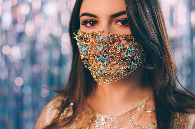 moda covid-19. Joyería festiva. Fiesta de Navidad en cuarentena. Retrato de una mujer morena sensual con una máscara facial de cadena de oro de estilo étnico sobre un fondo de oropel azul borroso y brillante.