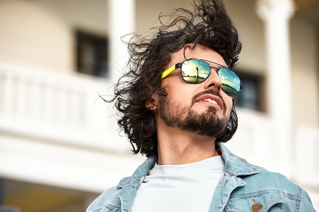 Moda de estilo de vida ao ar livre close-up retrato de cara muito bonito sorrindo e se divertindo Usando óculos de sol elegantes Em óculos de sol refletiu a paisagem da rua Cabelo escuro longo encaracolado e barba elegante