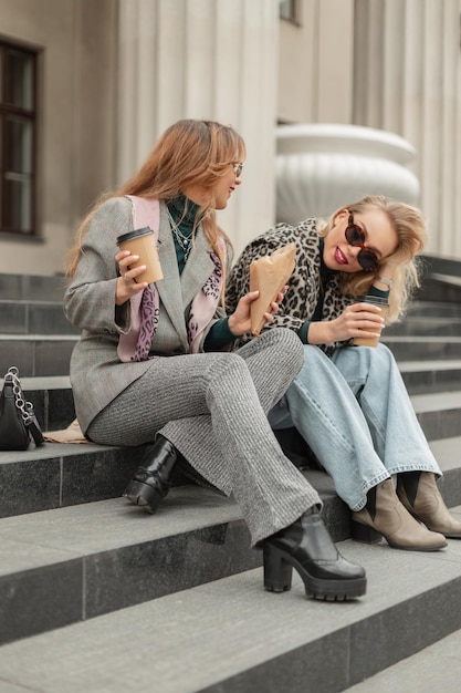 Moda divertida felicidad mujeres en ropa elegante de moda con zapatos se sienta en los escalones y bebe café en la ciudad Viajes y tiempo feliz