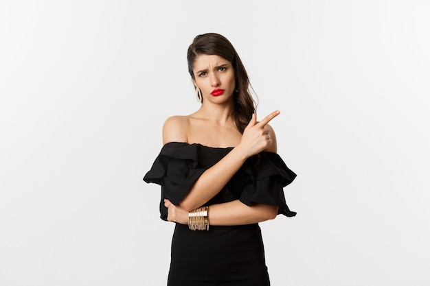 Moda e beleza. Mulher cética e glamourosa com lábios vermelhos, vestido preto, apontando o dedo direto para algo chato e chato, de pé sobre um fundo branco