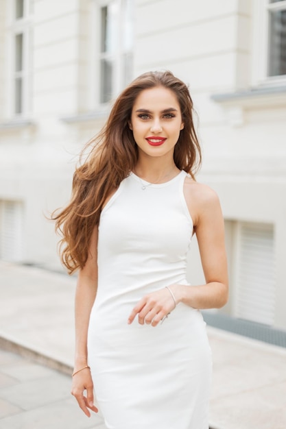 Moda elegante com sorriso com lábios vermelhos em vestido branco elegante caminha na rua perto de um edifício branco