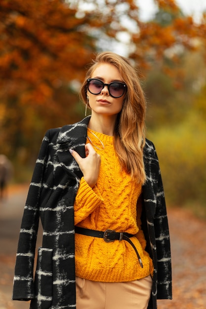 Moda elegante e linda mulher com óculos de sol estilosos em um suéter de malha amarelo vintage e casaco preto caminha na natureza de outono com folhas coloridas de ouro