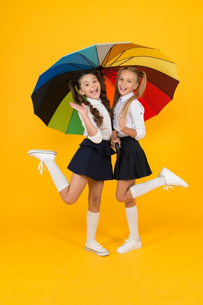 Moda de escuela primaria. Niños felices de la escuela con look de moda con paraguas de colores. Moda niñas pequeñas con uniformes formales sonriendo sobre fondo amarillo. Moda escolar para niños pequeños.