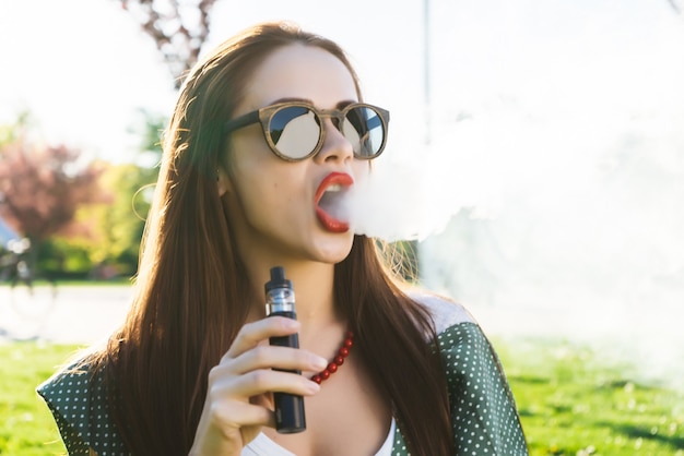 Foto moda feliz mujer sonriente en gafas de sol fumando vape en la calle, humo