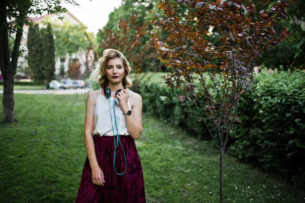 Moda y hermosa chica modelo rubia en elegante falda de terciopelo de terciopelo rojo, blusa blanca, posó en el parque con teléfono y auriculares.