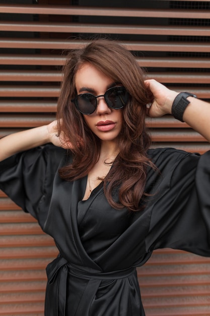 Moda hermosa joven modelo elegante con un peinado moreno con gafas elegantes en un elegante vestido negro en la calle cerca de una pared de metal