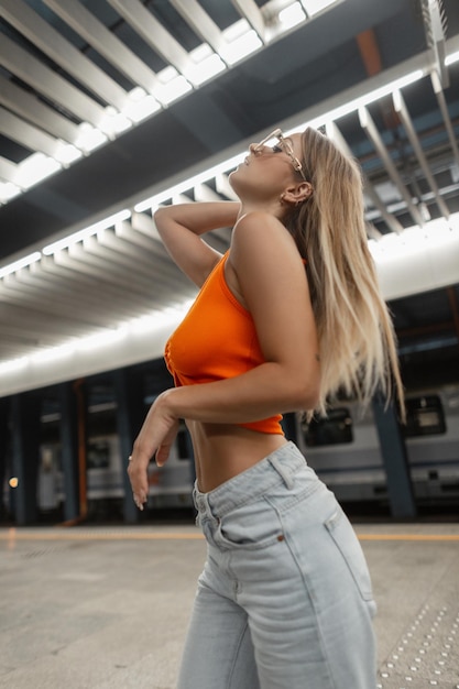 Moda hermosa modelo de mujer hipster urbana con gafas de sol blancas frescas con cuerpo en top naranja de moda con jeans vintage posando en el metro