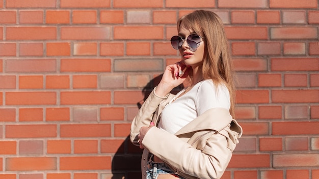 Moda hermosa mujer con estilo modelo hipster con gafas de sol redondas en ropa casual con estilo con chaqueta de cuero y top de pie cerca de la pared de ladrillo rojo