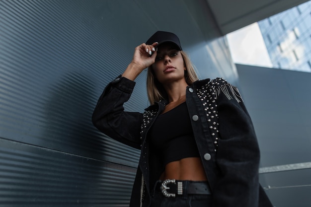 Moda hermosa mujer fresca con una gorra negra de maqueta de moda en una chaqueta de mezclilla glamorosa con pedrería posando en la ciudad cerca de un edificio moderno