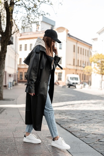 Moda hermosa mujer joven modelo hipster en ropa casual negra de moda con una gorra en un abrigo largo de cuero con jeans y zapatos blancos caminando en la ciudad Cool look urbano femenino