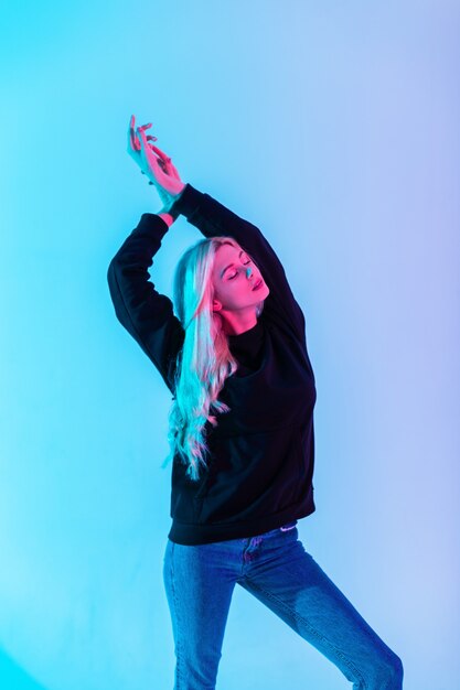 Moda hermosa mujer rubia sensual en sudadera con capucha negra con elegantes jeans azules en luz multicolor de neón rosa en estudio