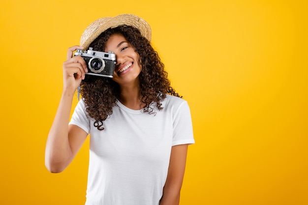 Moda hipster chica negra con cámara vintage y sombrero aislado sobre amarillo