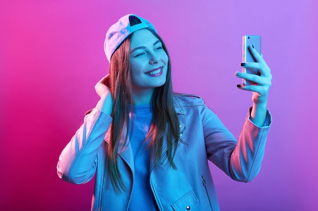 Foto moda hipster cool girl tomando autorretrato en un teléfono inteligente mientras posa aislado sobre el espacio de neón rosa