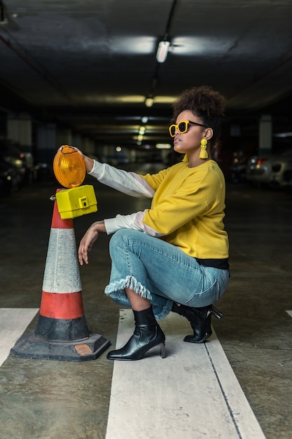 Moda hipster mujer negra en cuclillas cerca del cono de tráfico