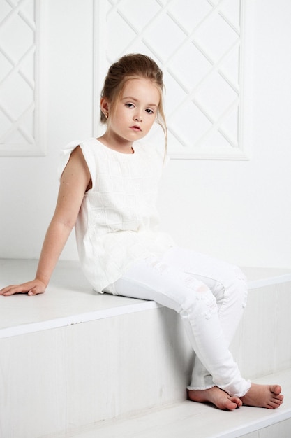 Moda infantil Hermosa chica rubia seria y pensativa vestida de blanco sentada y posando