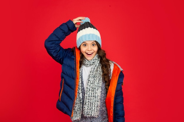 La moda de invierno sorprendió a un niño con el pelo rizado en una chaqueta acolchada y un sombrero. Una adolescente con un retrato de fondo rojo de un niño con ropa abrigada y una bufanda expresa una emoción positiva.