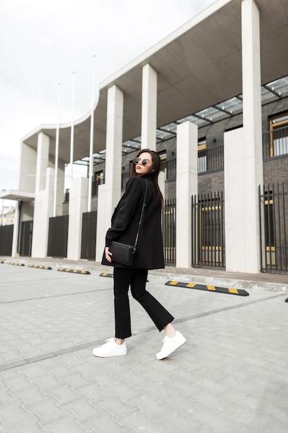 Moda jovem adorável com bolsa de couro em elegantes óculos de sol em elegantes roupas pretas da nova coleção da moda jovem caminha perto de um edifício moderno na cidade. Garota legal urbana posa na rua.
