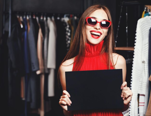 Foto moda jovem bem sucedida usando óculos escuros e vestidos vermelhos mostrando cartão preto no shopping de moda durante o processo de compras conceito de consumismo black friday venda vida rica